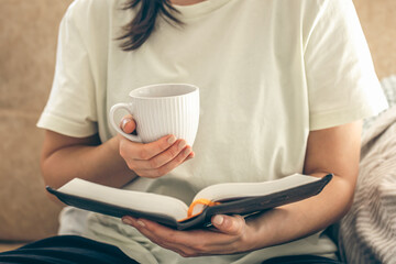 Woman holding a cup of tea and reading a book on the sofa at home, close up.