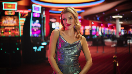 Fototapeta premium Beautiful Young Caucasian Woman Posing In Prestige Casino Next To Baccarat Table. Charismatic Female Croupier Smiling, Looking At Camera And Wearing Fancy Silver Dress