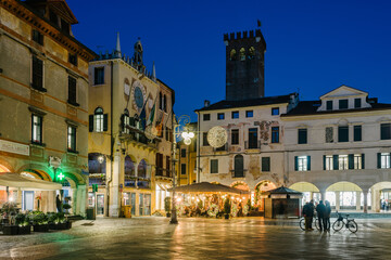 Bassano del Grappa, Italy - January 09, 2024: People Piazza Libertà in cafe tables in the evening in winter. On the left the town hall. - 706457196