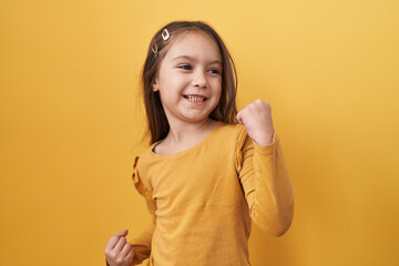 Adorable hispanic girl joyfully standing, arms raised in victorious celebration of success on a yellow isolated background, eyes softly closed, yes!