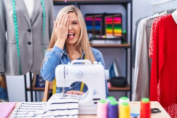 Blonde woman dressmaker designer using sew machine covering one eye with hand, confident smile on face and surprise emotion.