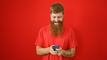 Handsome young redhead man, brimming with happiness, confidently typing away messages on his smartphone over red isolated background. his laughter and casual positivity are infectious.