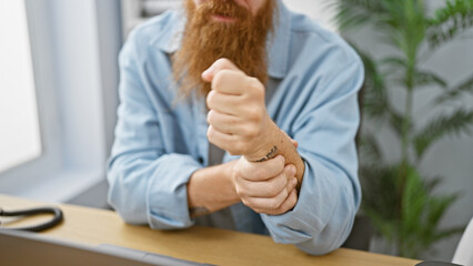 Overworked handsome young redhead man stressed with wrist pain from laptop work at busy indoor...