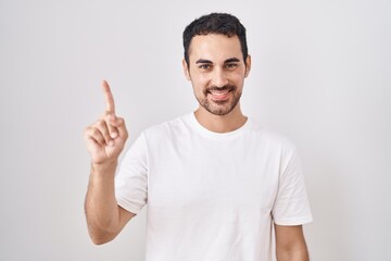Handsome hispanic man standing over white background showing and pointing up with finger number one...