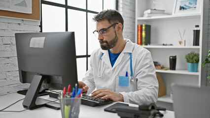 Bearded young hispanic man doctor hard at work, using his computer indoors at a medical clinic