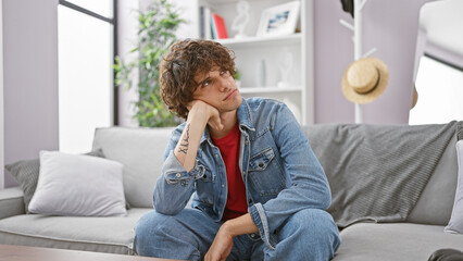 Pensive young man with curly hair, wearing denim, sitting on a couch in a modern living room. - obrazy, fototapety, plakaty
