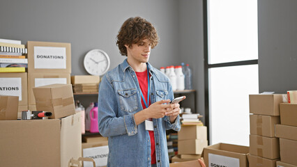 Young man texting on smartphone amidst boxes inside a donation center.