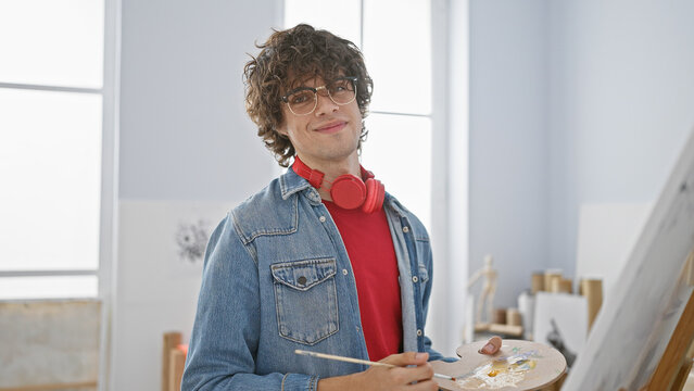 Confident young man with glasses and headphones stands in an art studio, holding a paint palette and brush.