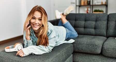 Young caucasian woman talking on the smartphone lying on sofa at home