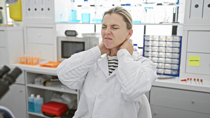 A young caucasian woman in a lab coat winces in pain, touching her neck in a laboratory setting,...
