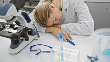 Fototapeta premium A tired woman rests her head on a lab table next to a microscope and test tubes, depicting exhaustion in a scientific setting.