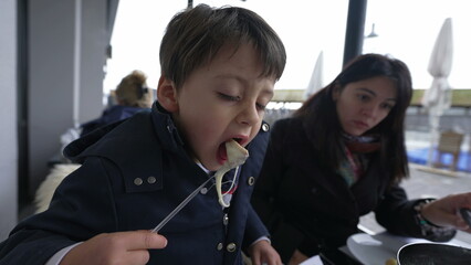 Child blowing hot food while eating traditional Swiss fondue at restaurant with mom. Parent and...