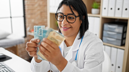Smiling woman doctor holding brazilian money in a clinic office, implying healthcare costs or...