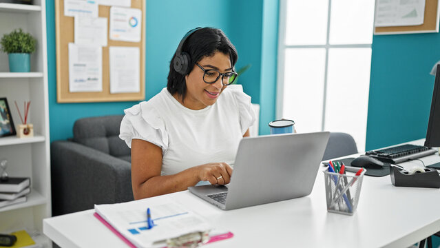 A smiling woman with headphones using a laptop in a modern office with turquoise walls and organized desk.