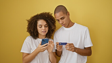 Beautiful couple in love, engrossed in online shopping with their smartphone and credit card, standing against a isolated yellow background