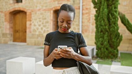 Fototapeta premium Beautiful african american woman shares a happy smile while using mobile phone in a lush city park