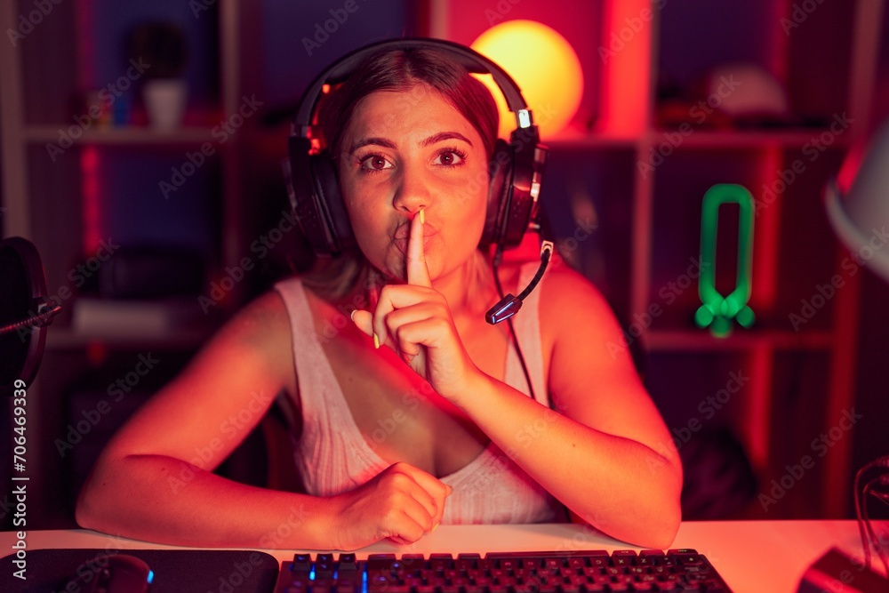 Poster Young blonde woman playing video games wearing headphones asking to be quiet with finger on lips. silence and secret concept.