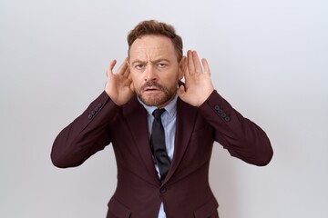 Middle age business man with beard wearing suit and tie trying to hear both hands on ear gesture, curious for gossip. hearing problem, deaf