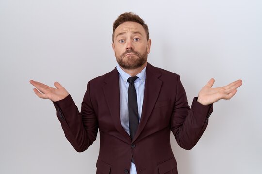 Middle age business man with beard wearing suit and tie clueless and confused expression with arms and hands raised. doubt concept.