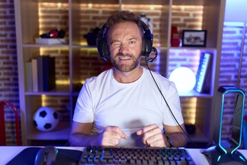 Middle age man with beard playing video games wearing headphones winking looking at the camera with...