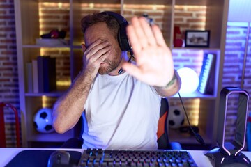 Middle age man with beard playing video games wearing headphones covering eyes with hands and doing stop gesture with sad and fear expression. embarrassed and negative concept.