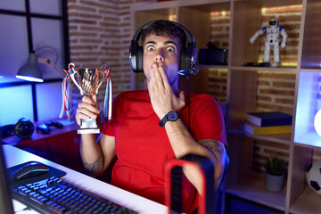 Young hispanic man playing video games holding trophy covering mouth with hand, shocked and afraid for mistake. surprised expression