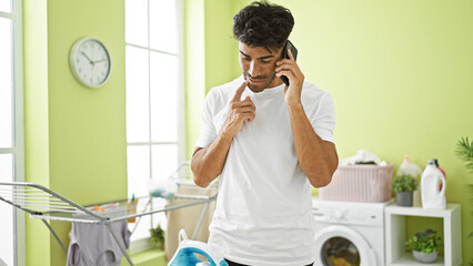 Hispanic man talking on phone in a bright laundry room, wearing a white shirt, looking...