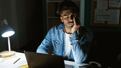 Tired hispanic man working late in office, illuminated by lamp with a laptop and documents.