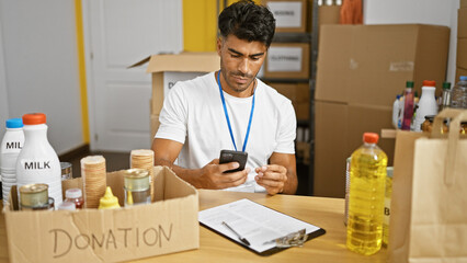 Handsome hispanic man checks phone at donation center surrounded by food and boxes.