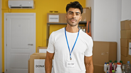 Handsome young hispanic man with beard wearing a volunteer badge stands confidently in a storage...
