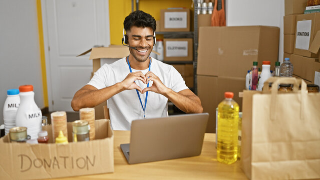 A cheerful man forms a heart with his hands in a warehouse with donation boxes, food, and laptop signaling gratitude and positivity.