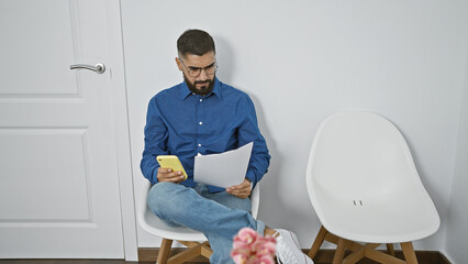 Bearded man in blue shirt reviewing documents and using smartphone indoors.