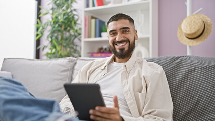 A smiling man with a beard holds a tablet while relaxing on a couch in a cozy living room.