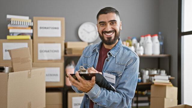 Handsome man with beard volunteering in a warehouse, writing on clipboard amid donation boxes.
