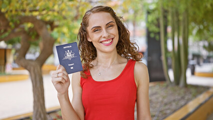 Confident young woman joyfully smiling, proudly holding her american passport in the green park,...