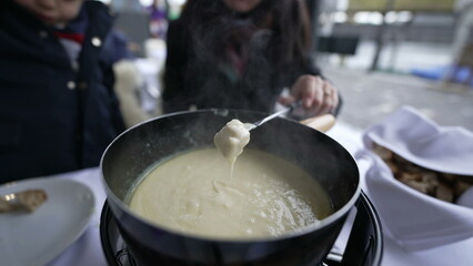 Closeup of traditional Swiss fondue, people eating bread with cheese at outdoor restaurant enjoying...