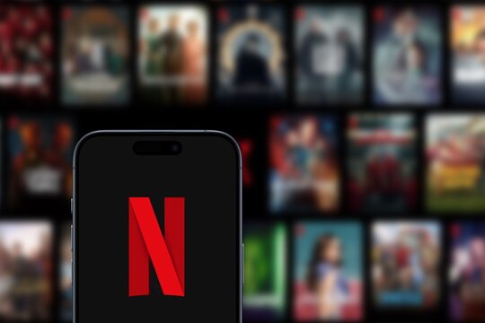 Netflix logo is displayed on a modern smartphone, with a selection of series and films from the streaming platform displayed in the background, 2024