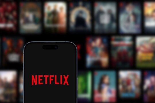 Netflix logo is displayed on a modern smartphone, with a selection of series and films from the streaming platform displayed in the background, 2024