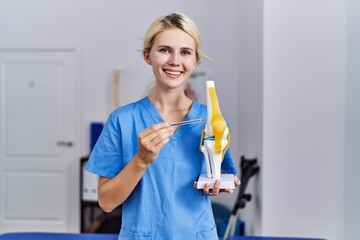 Young blonde woman pysiotherapist smiling confident pointing to anatomical model of knee at rehab clinic
