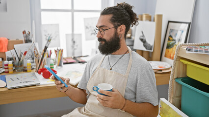 Hispanic man with beard using phone in art studio while holding a coffee cup.