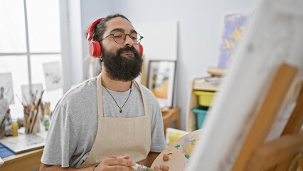 A young hispanic man wearing headphones paints on a canvas in a bright art studio.