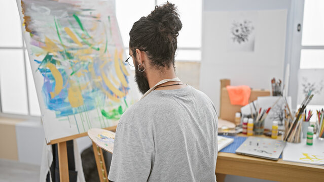 Handsome hispanic man with beard considering a colorful painting in an art studio setting.