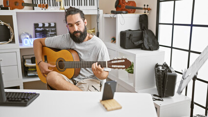 Handsome bearded hispanic man playing guitar in a modern music studio with instruments.