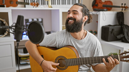 A smiling bearded man plays an acoustic guitar in a home music studio with microphones and...