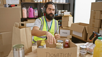 Bearded man wearing headphones packages food donations in a busy warehouse.