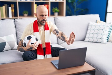 Hispanic man with tattoos watching football match hooligan holding ball on the laptop with open...