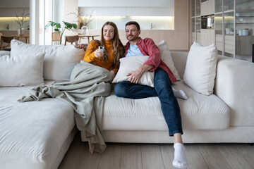Married couple watch TV together sitting on couch at home enjoying favourite movie series. Pleased woman holding remote control with loving man, ready to change television channel. Family lazy weekend - Powered by Adobe