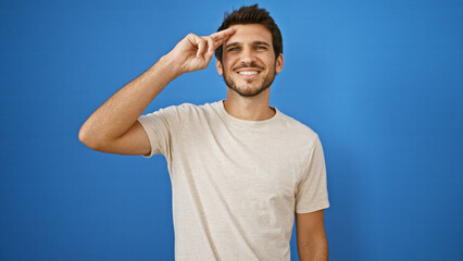 Handsome young hispanic man saluting isolated on a blue wall background outdoor