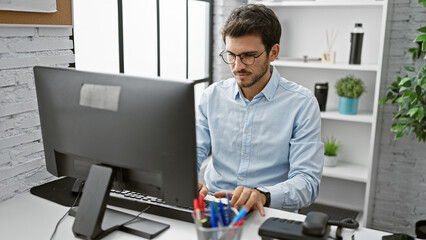 A young hispanic man in a blue shirt working attentively at his office desk indoors, illustrating a...