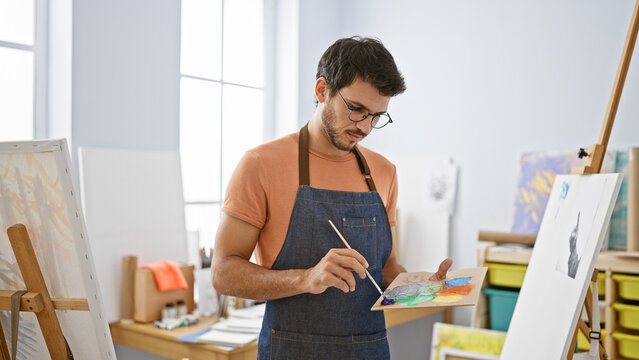 A focused man with glasses and beard painting in a bright art studio.